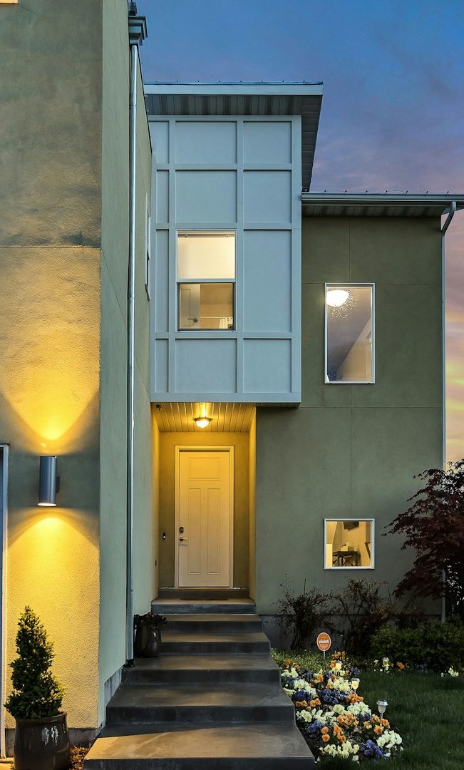 Modernes Haus mit Eingangstür, sanften Beleuchtungen und blühenden Blumen.